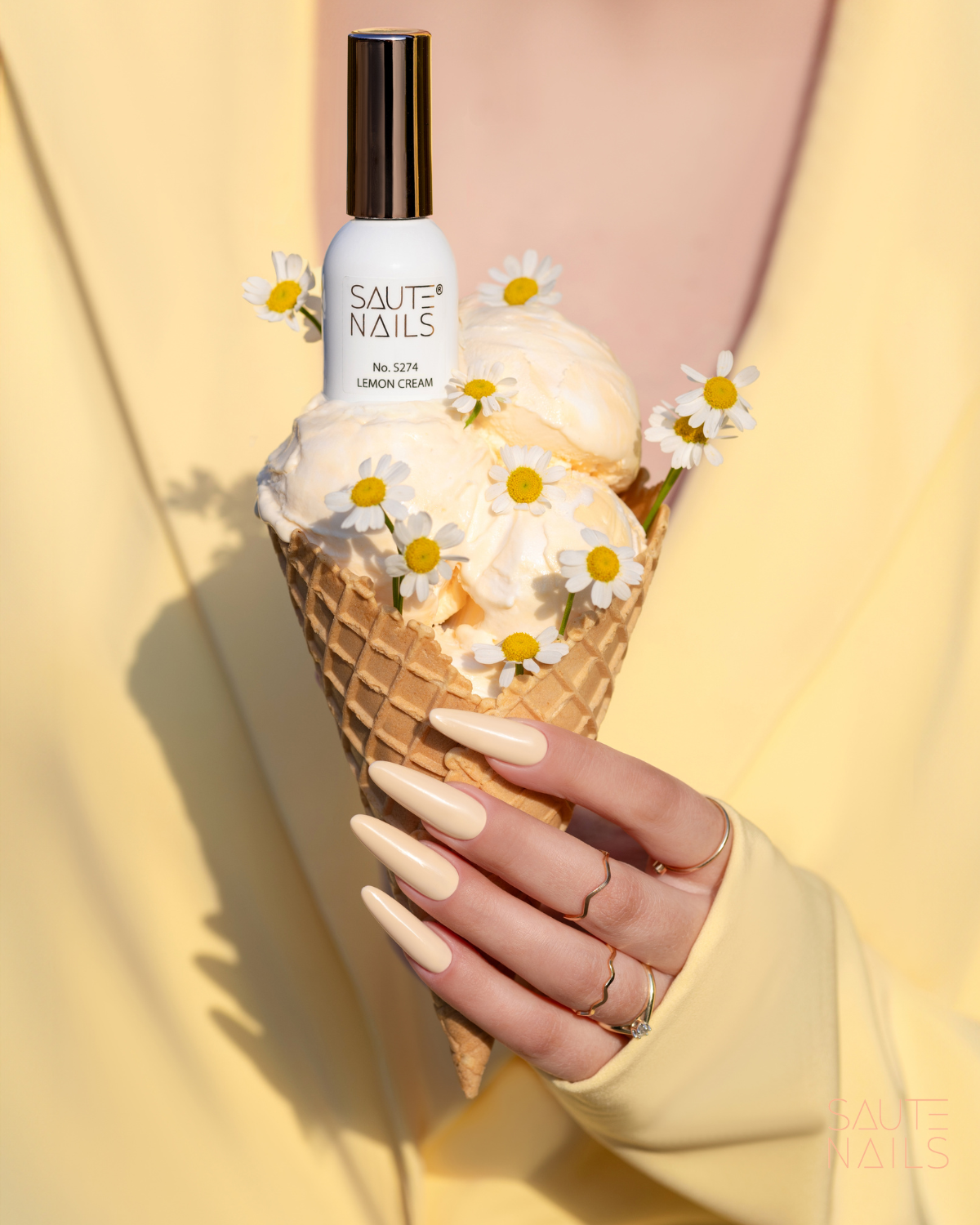 Verniz Gel Saute Nails S274 - Lemon Cream (Amarelo Cremoso) Frasco de verniz SAUTE NAILS num cone de gelado com creme e flores, mão com unhas creme a segurar.