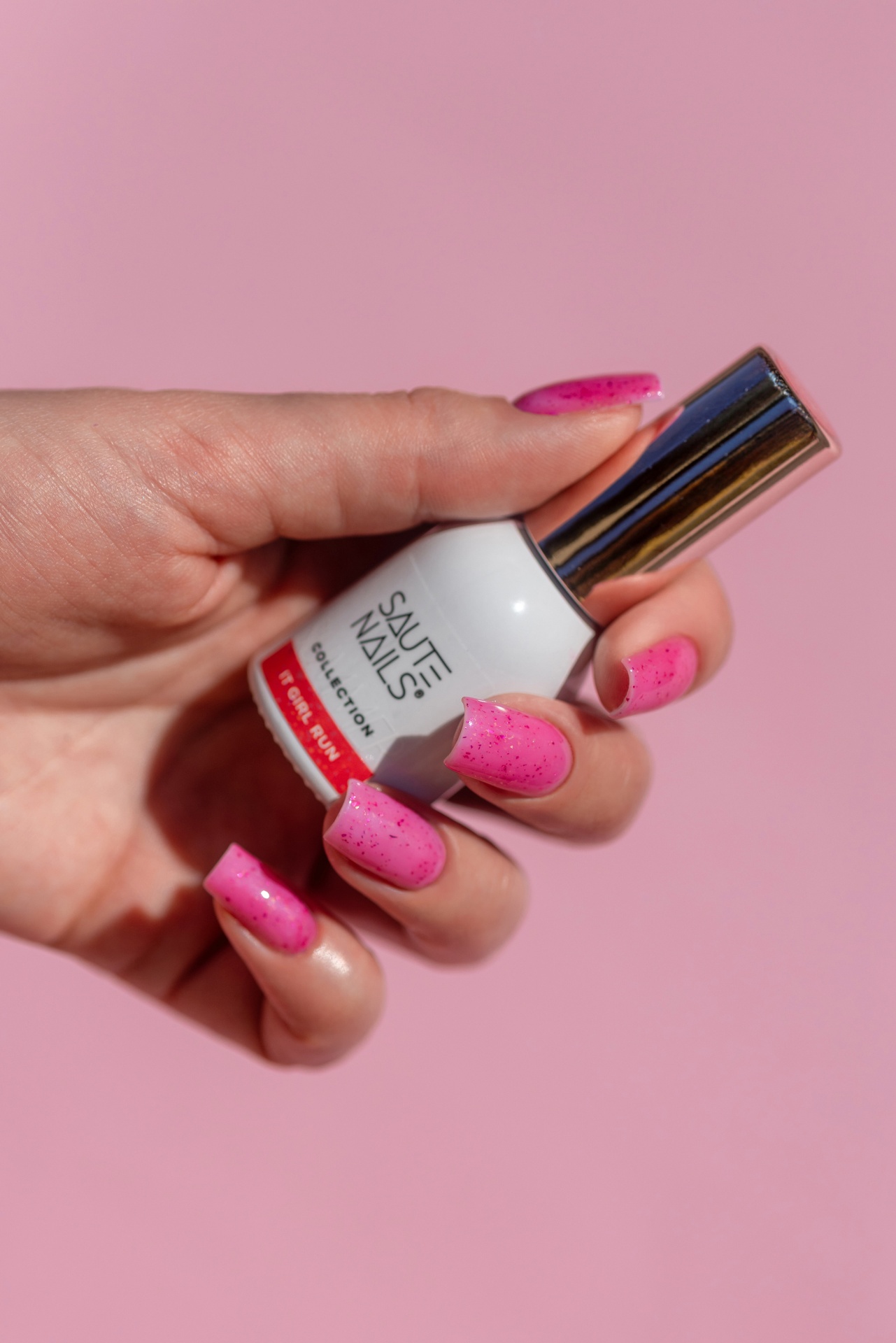 Verniz Gel It Girl Run (Rosa Néon com partículas Rosa Escuro) Mão feminia segurando frasco de verniz SAUTÉ NAILS branco com tampa metálica