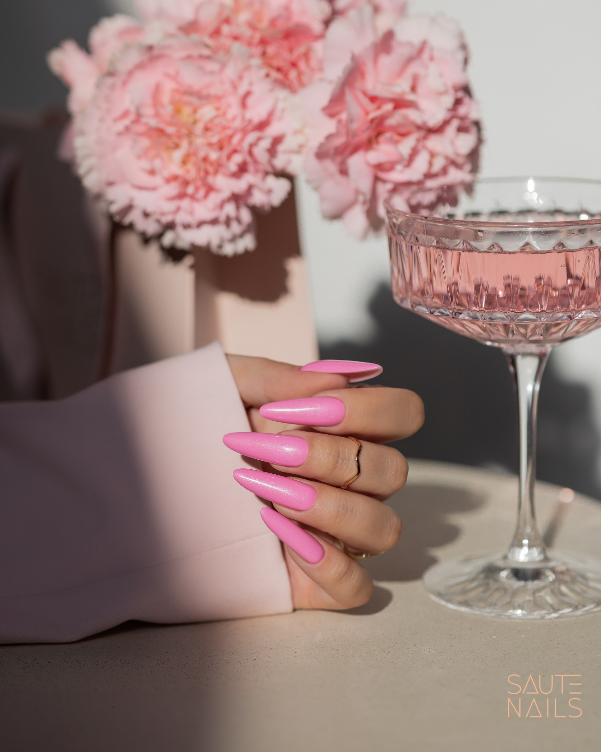 Verniz Gel Saute Nails S273 - Sparkling Spring (Rosa com Glitter) Mão com unhas longas cor-de-rosa junto a taça de vidro e flores cor-de-rosa