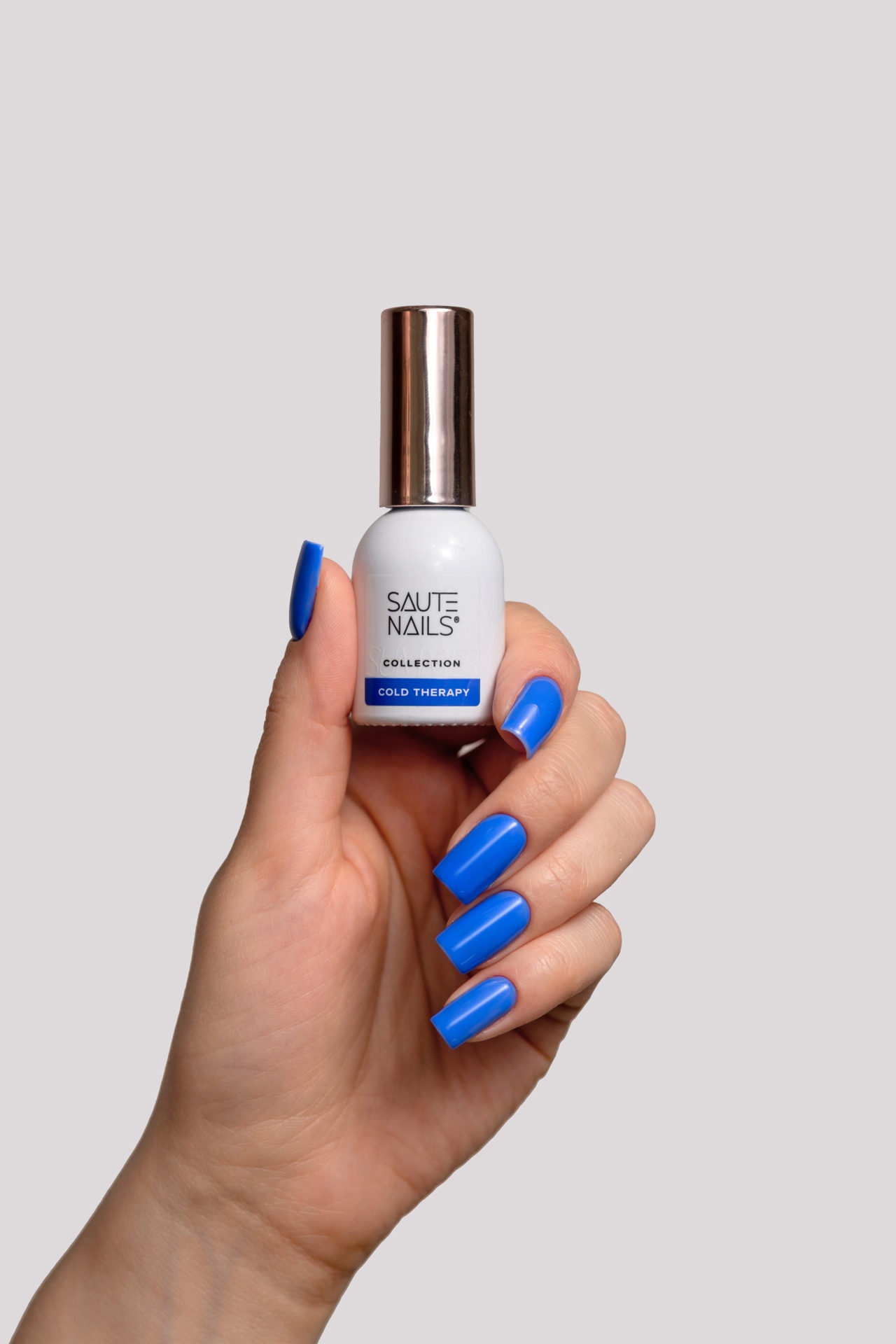 Verniz Gel Cold Therapy (Azul Royal Intenso) Frasco branco de verniz para unhas com tampa metalizada, segurado por mão com unhas azuis
