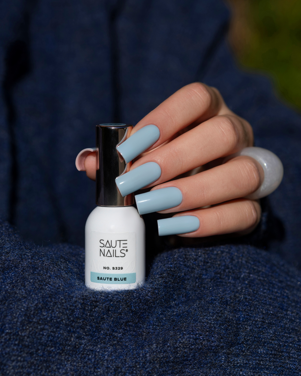Verniz Gel Saute Nails S329 - Saute Blue (Azul Suave) Frasco de verniz de unhas branco com tampa prateada e unhas pintadas de azul claro