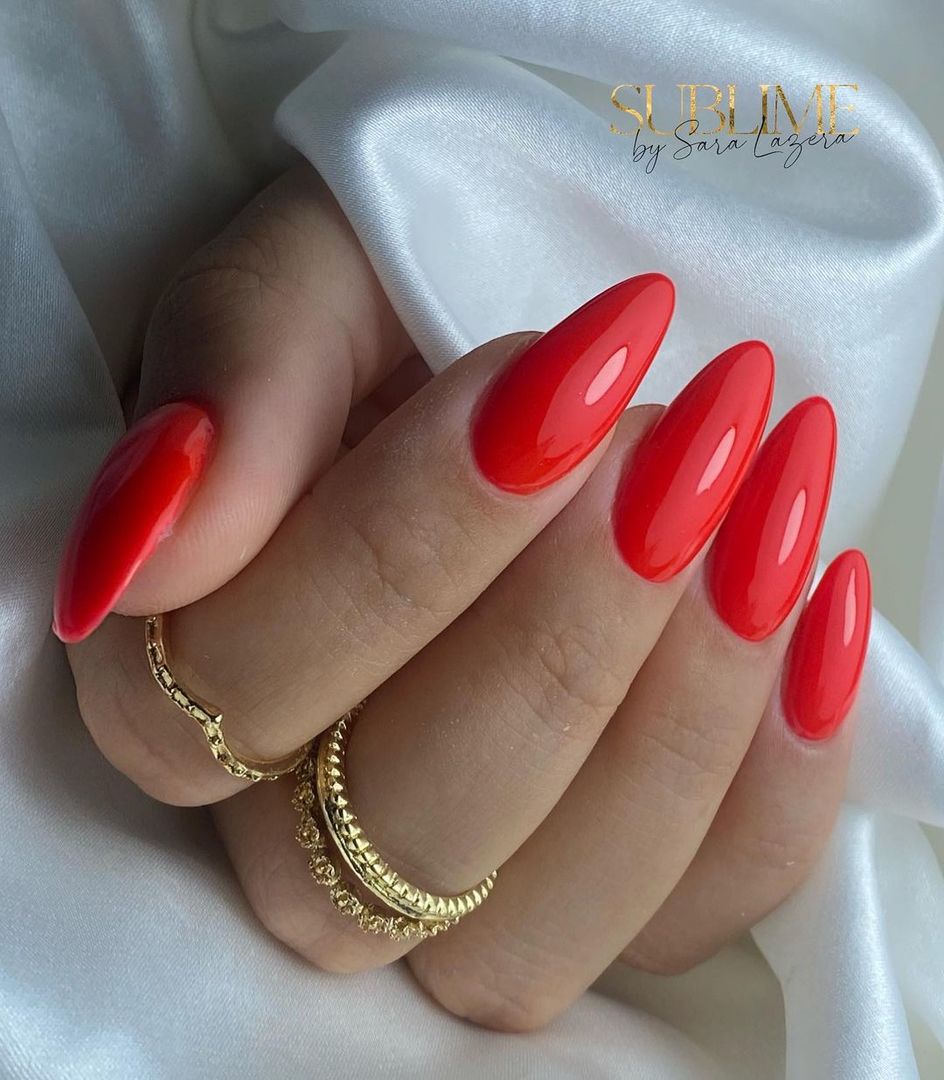 Verniz Gel Andreia 293 (Vermelho Coral) Mão com unhas vermelhas brilhantes e anéis dourados sobre tecido branco