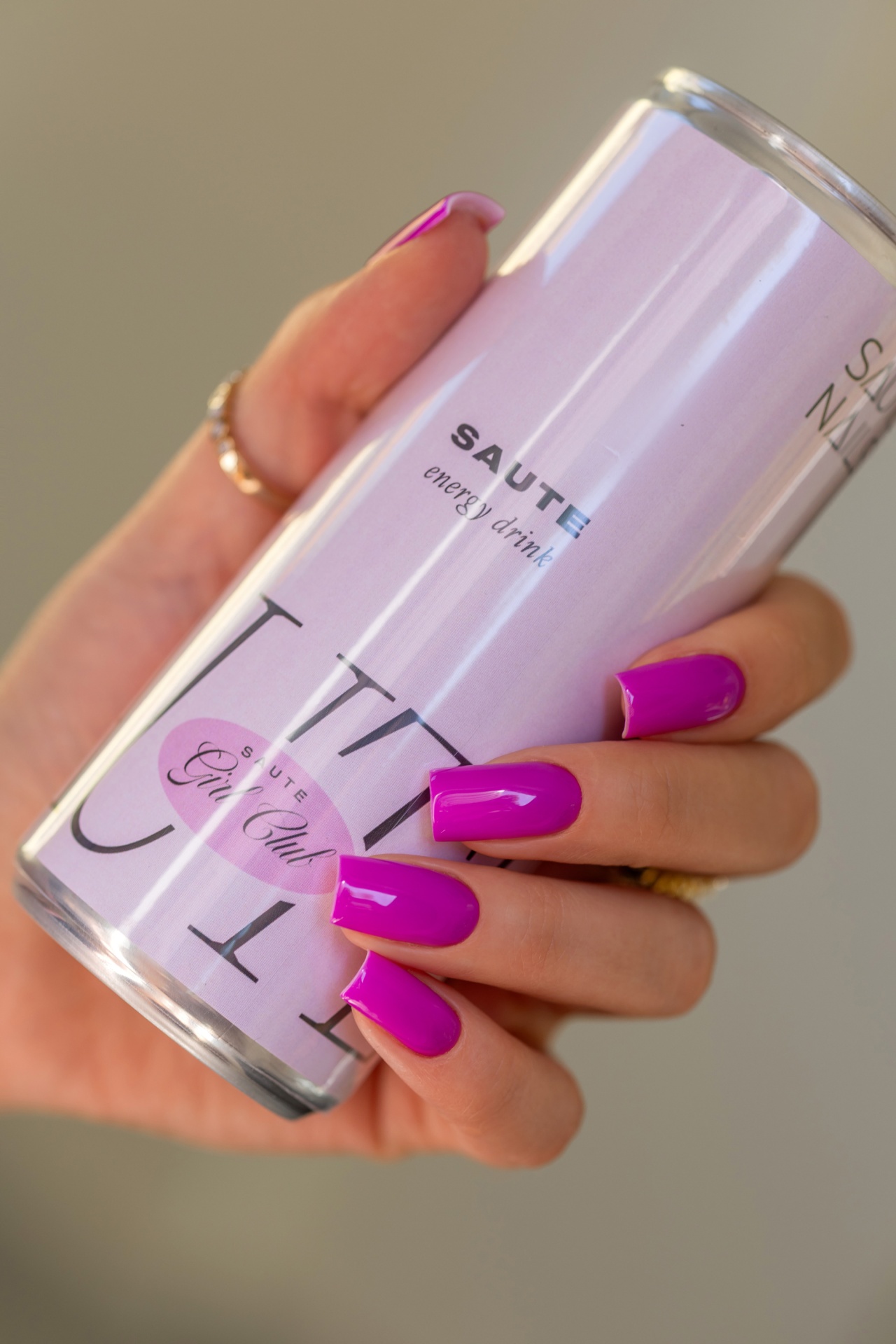 Verniz Gel Hot Pilates Club (Fúcsia‑Violeta Néon) Mão com unhas roxas segurando uma lata lilás de bebida energética.