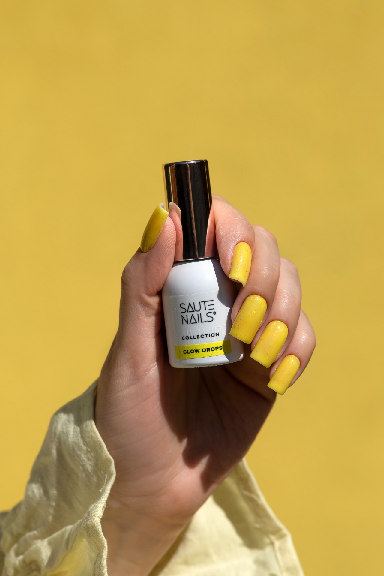 Verniz Gel Glow Drops (Amarelo Intenso com Partículas Douradas) Frasco de verniz SAUTE NAILS branco com unhas amarelas brilhantes