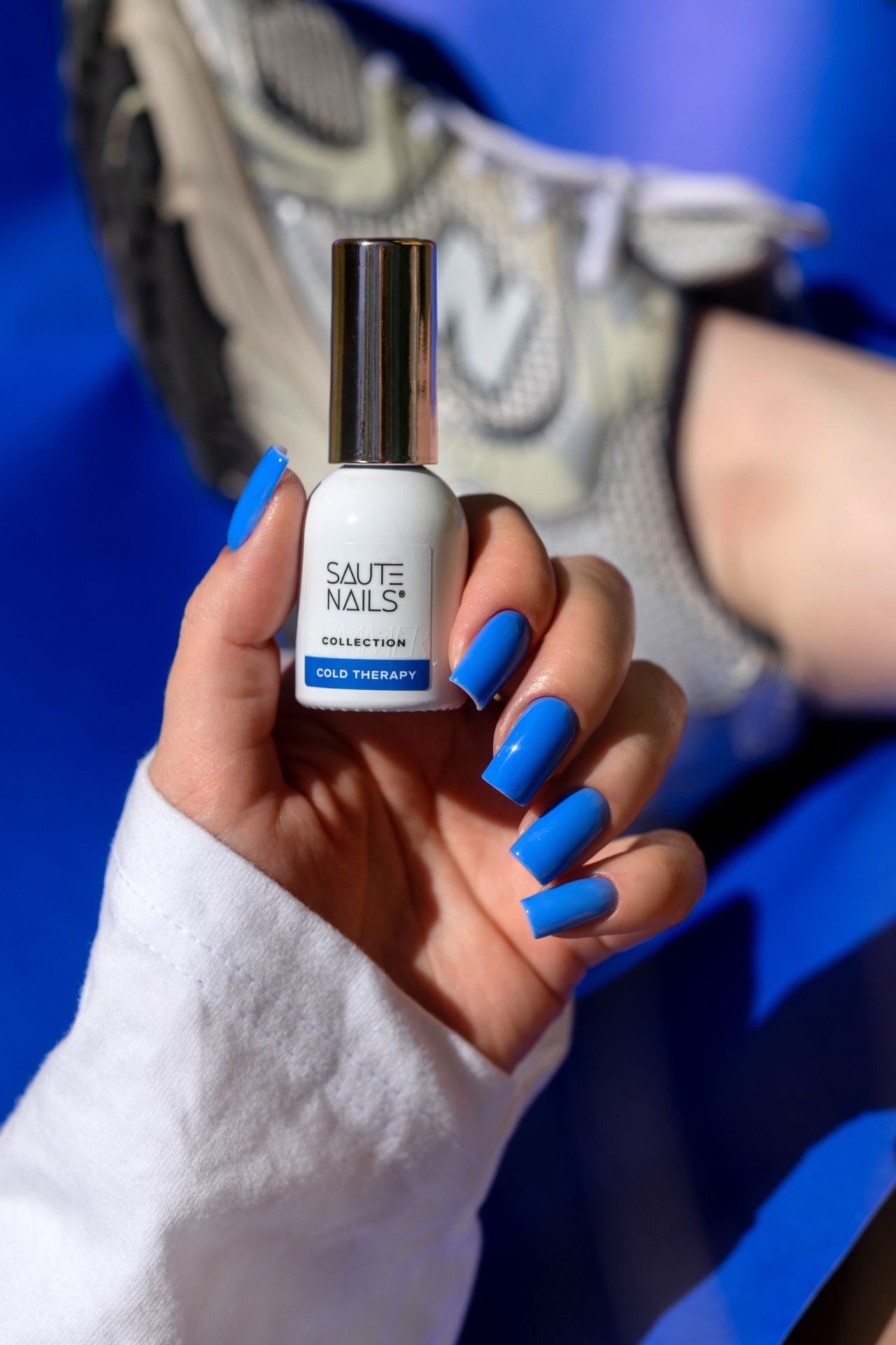 Verniz Gel Cold Therapy (Azul Royal Intenso) Frasco de verniz para unhas branco com tampa metálica e unha pintada de azul