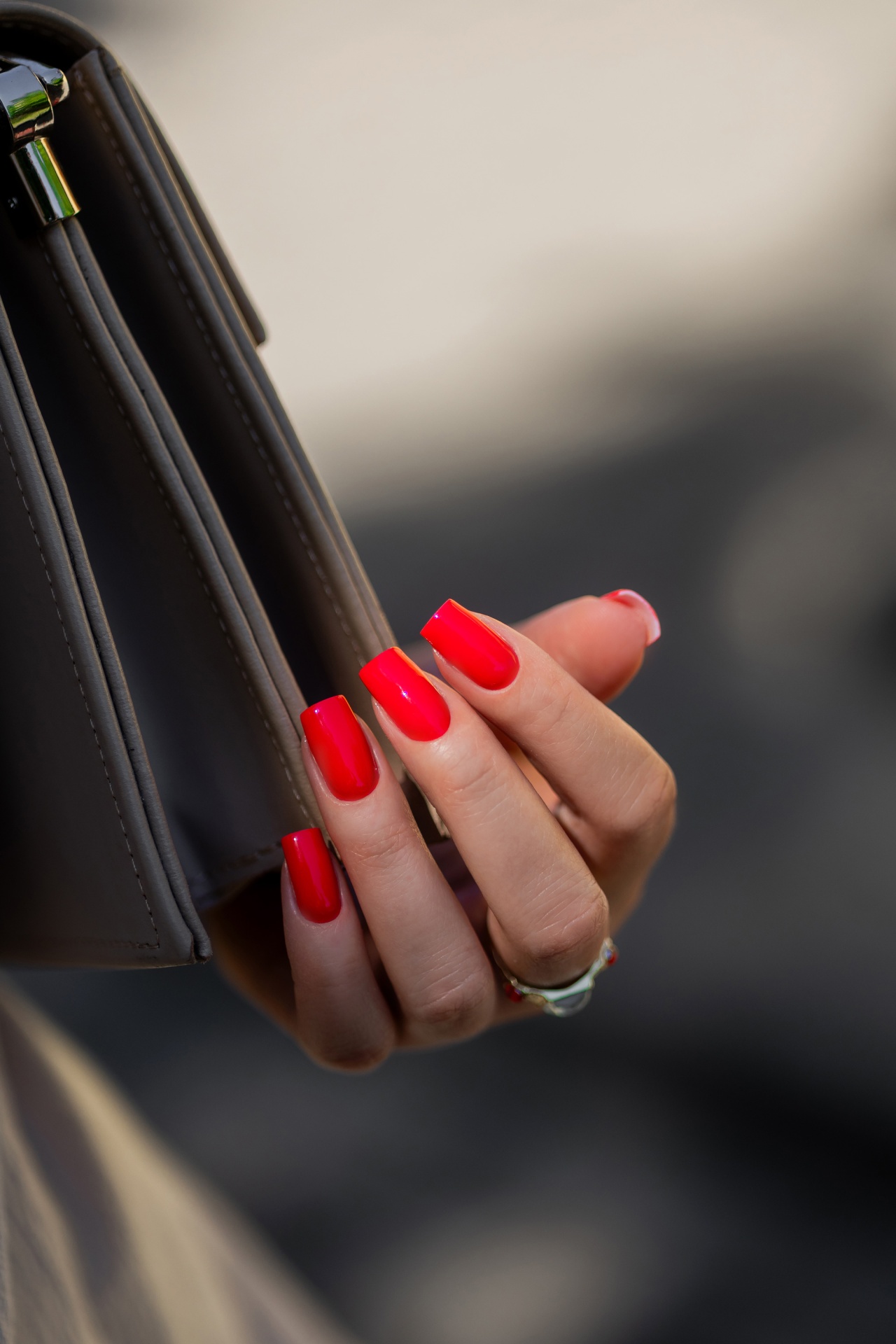 Verniz Gel Red Light Baby (Vermelho Laranja Néon) Mão com unhas vermelhas segurando mala de couro cinzento