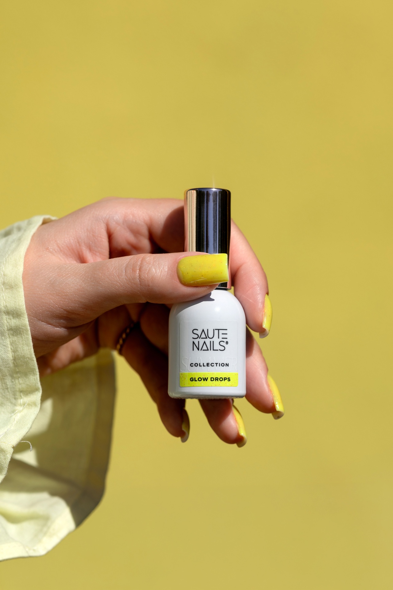 Verniz Gel Glow Drops (Amarelo Intenso com Partículas Douradas) Frasco branco de verniz de unhas com tampa prateada e mãos com unhas amarelas a segurá-lo