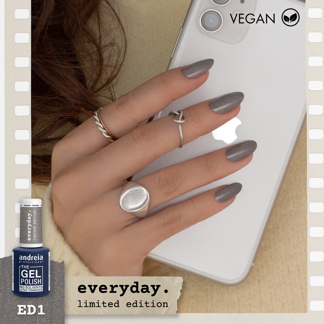 The Gel Polish Andreia ED1 - Cinza com shimmer dourado Mão com unhas prateadas e anéis segurando smartphone branco, ao lado verniz gel andreia azul escuro, texto vegan e everyday limited edition