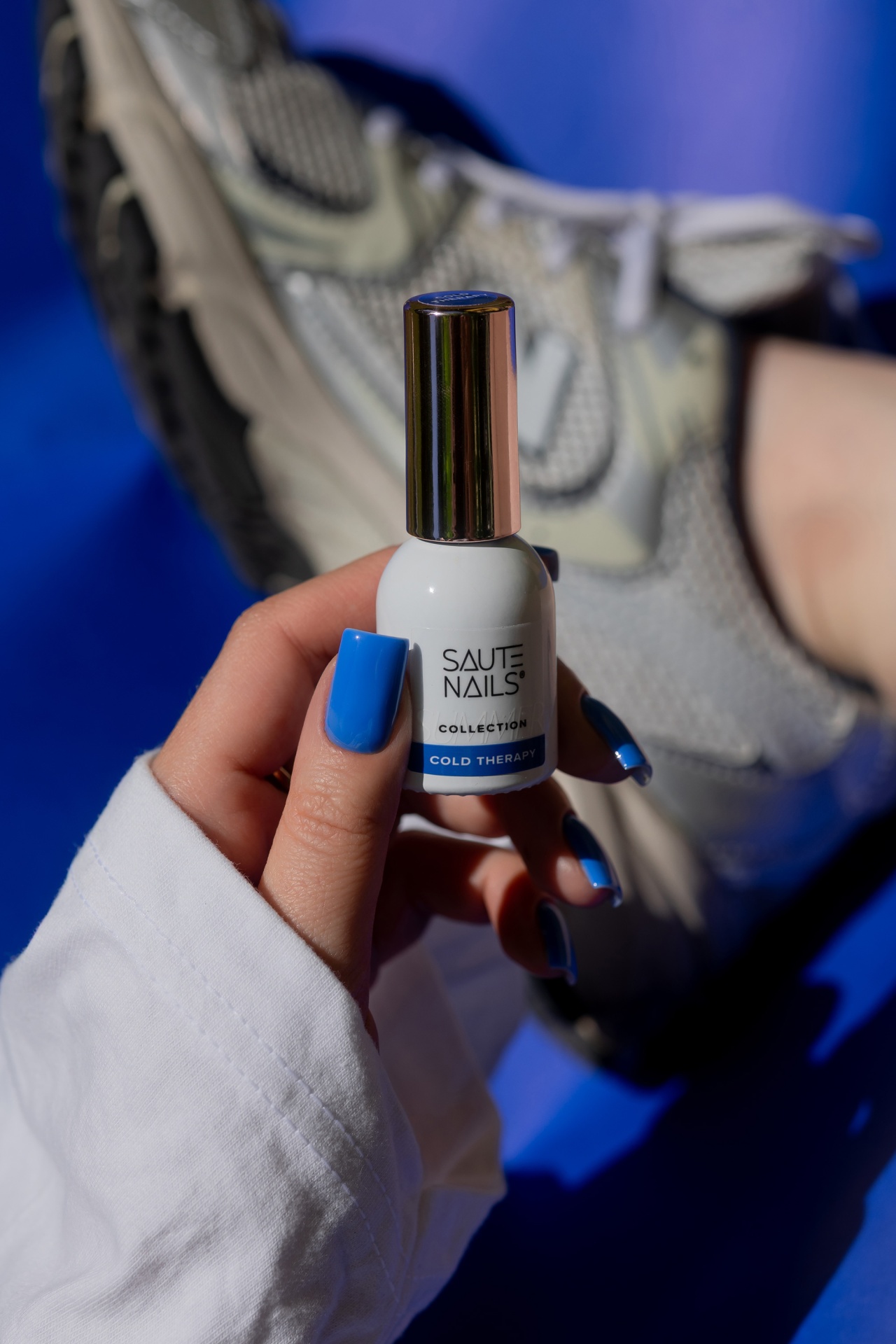 Verniz Gel Cold Therapy (Azul Royal Intenso) Frasco de verniz para unhas branco com tampa metálica e texto, unhas azuis, ténis cinzentos ao fundo.