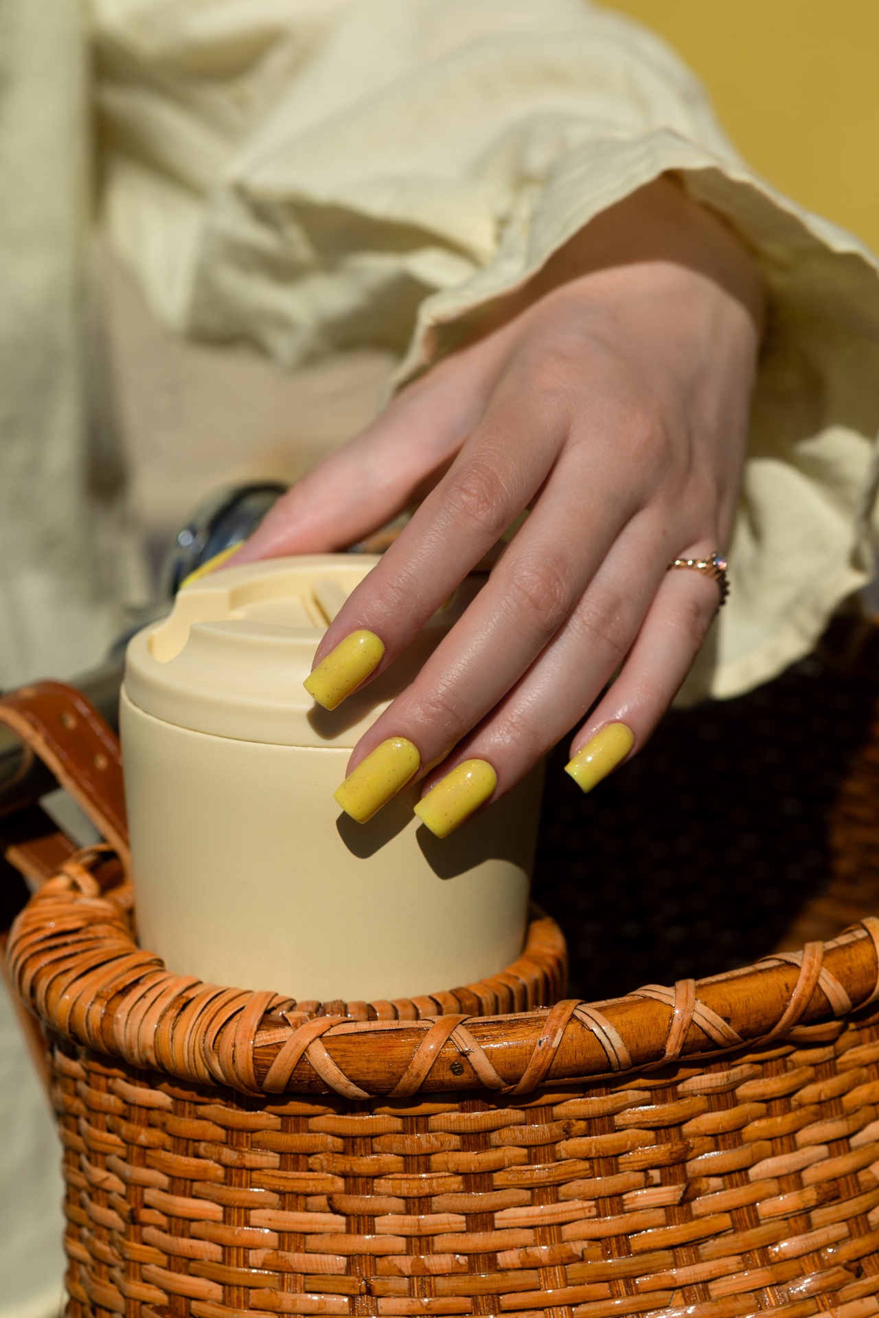 Verniz Gel Glow Drops (Amarelo Intenso com Partículas Douradas) Mão feminina com unhas amarelas segurando copo térmico bege num cesto de verga