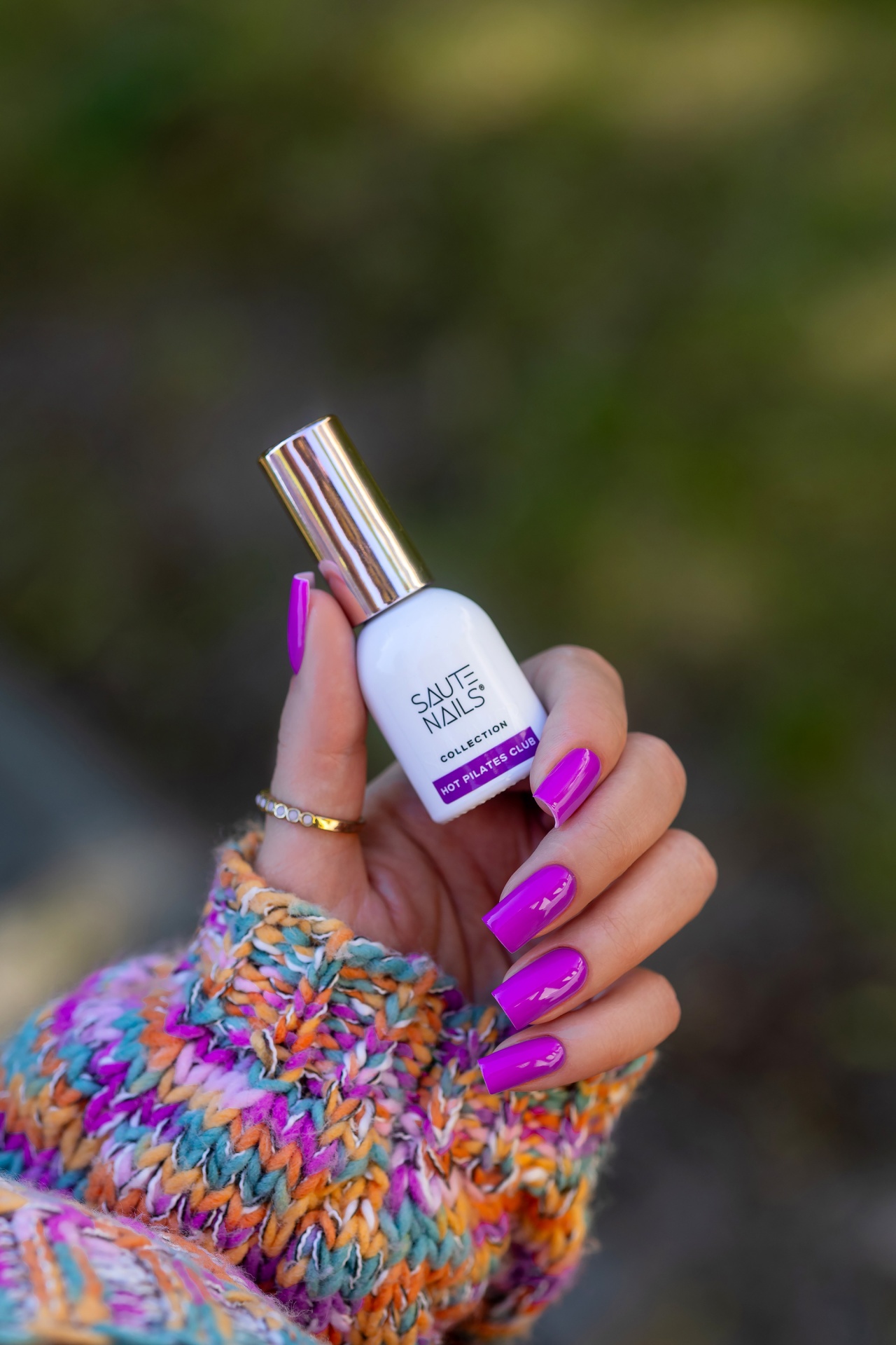 Verniz Gel Hot Pilates Club (Fúcsia‑Violeta Néon) Frasco branco de verniz para unhas com tampa dourada, segurado por mão com unhas roxas e camisola colorida.