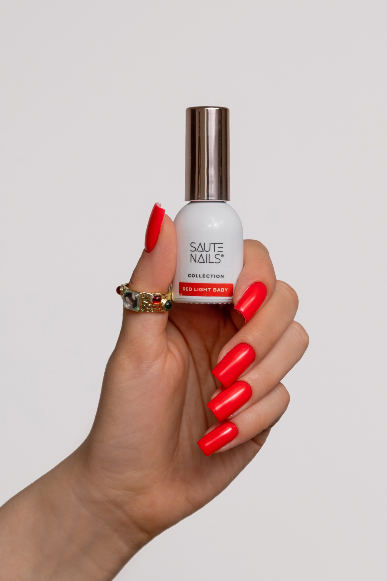 Verniz Gel Red Light Baby (Vermelho Laranja Néon) Frasco de verniz de unhas branco com tampa prateada segurado por mão com unhas vermelhas e anel dourado