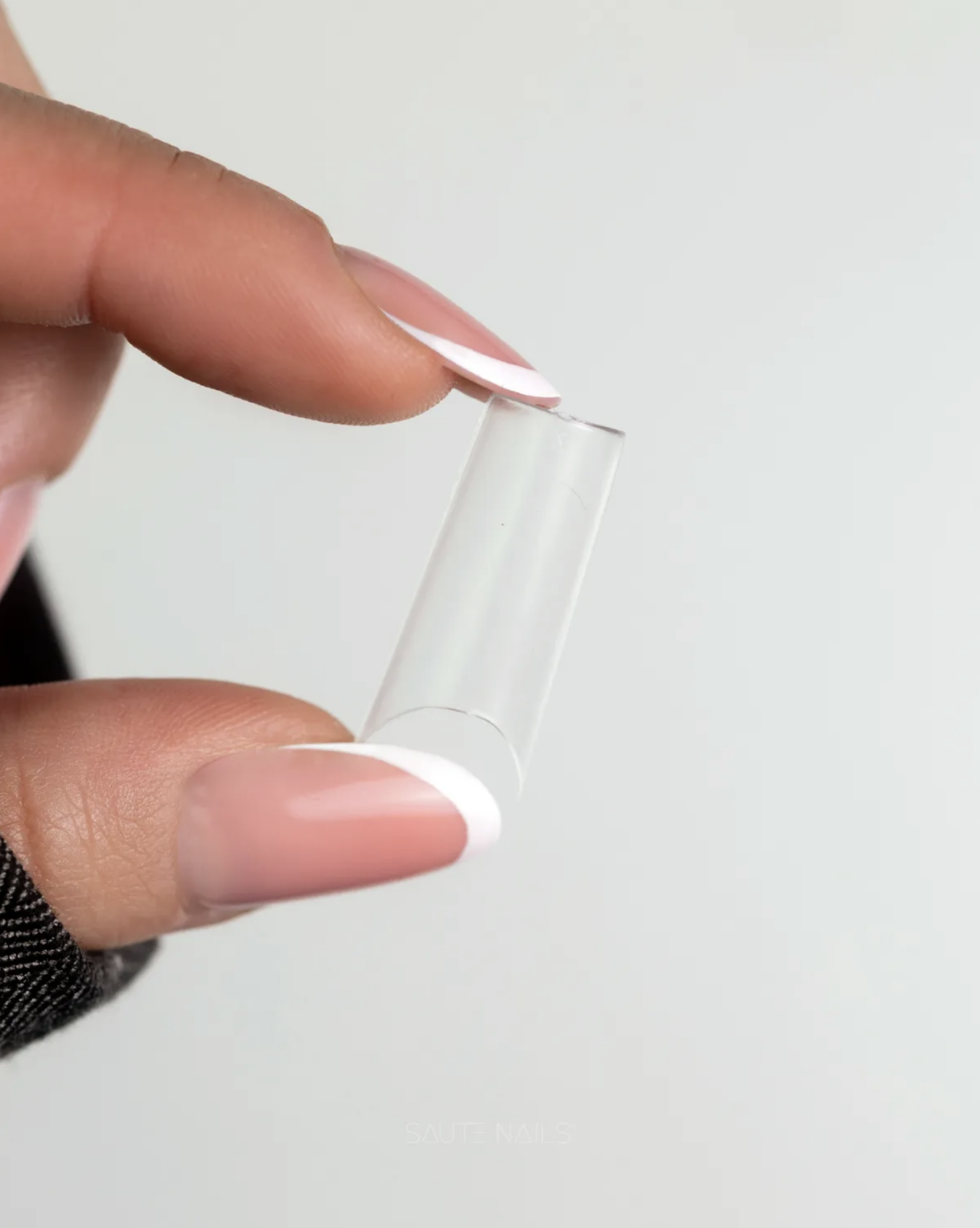 Tunnel Tips Ultra-Finas Saute Nails - 504 unidades Unha falsa transparente sendo segurada entre mãos com unhas pintadas de rosa claro e branca