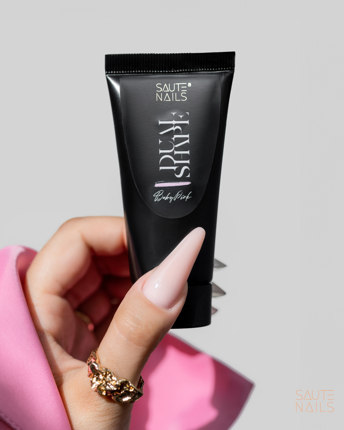 Saute Nails Dual Shape Baby Pink 35g Mão com unha longa rosa claro segurando tubo preto de plástico com texto branco Saute Nails Poly Shape Baby Pink