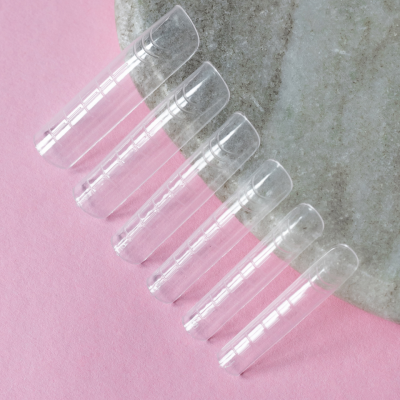 Seis unhas postiças transparentes em tamanhos variados sobre fundo rosa e pedra cinzenta