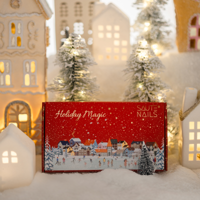 Caixa de Natal vermelha Holiday Magic da SAUTE NAILS em cenário de Natal com casas e árvores nevadas