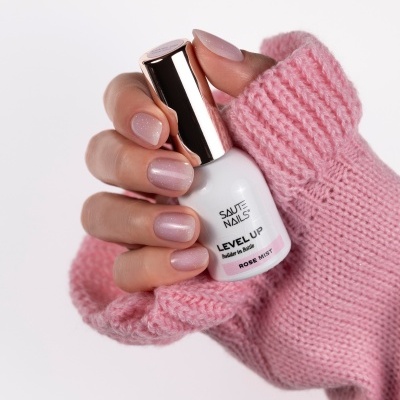 Mão com unhas pintadas de rosa claro brilhante segurando verniz SAUTE NAILS LEVEL UP ROSE MIST com camisola rosa.