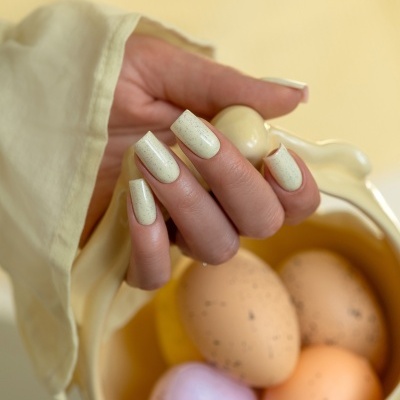 Mão feminina com unhas bege claro manchadas segurando recipiente amarelo com ovos coloridos manchados.