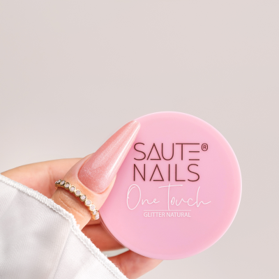 Frasco rosa com texto SAUTE NAILS One touch GLITTER NATURAL na mão com unha rosa brilhante e anel