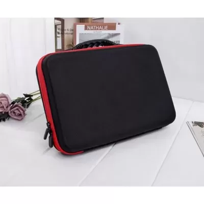 Estojo rígido preto com fecho vermelho e alça, sobre superfície branca com flores e livro ao fundo