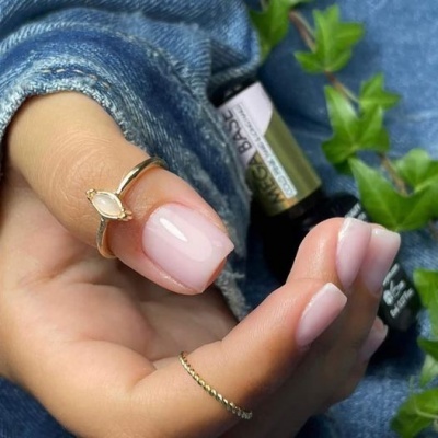 Mão com unhas rosa suave e anéis dourados, garrafa e folhas ao fundo
