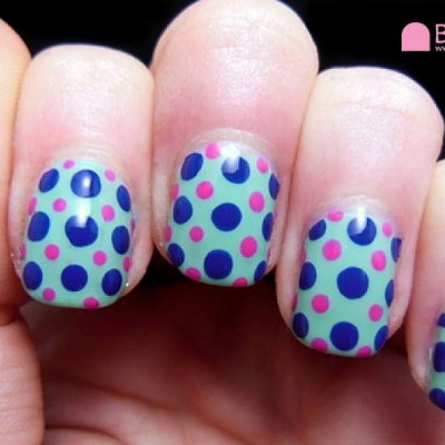 Unhas com verniz azul claro e pintas azuis e rosa em mão contra fundo preto