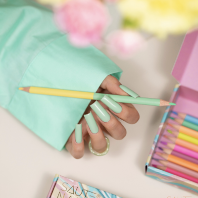 Mão com unhas verdes segurando lápis de cor ao lado de caixa com lápis coloridos