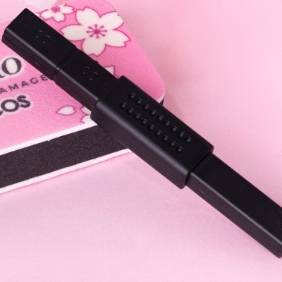 Produto preto com textura em pontos sobre superfície rosa com cartão floral e texto parcial