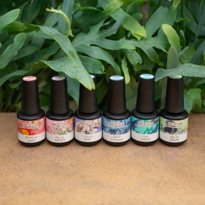 Seis frascos de verniz para unhas INOCOS com rótulos coloridos em superfície castanha perante folhas verdes