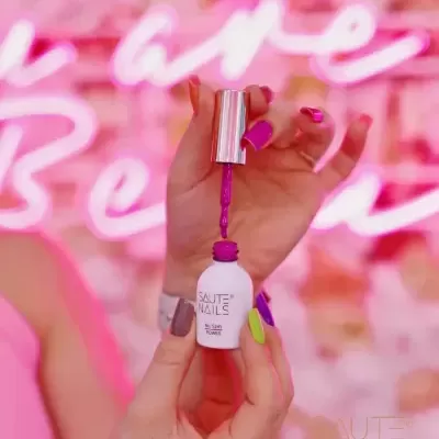 Frasco branco de verniz de unhas SAUTE NAILS sendo aplicado em unhas coloridas com fundo rosa neon