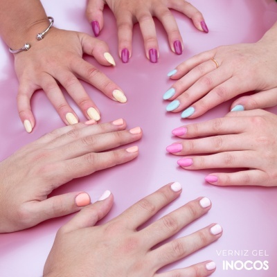 Mãos femininas com unhas pintadas em verniz gel colorido de várias cores pastel e vibrantes sobre fundo rosa.