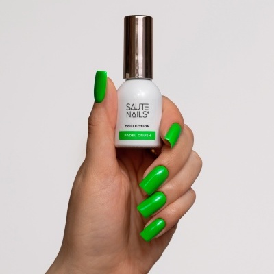 Frasco de verniz de unhas branco com tampa prateada e texto em verde e preto, em mão com unhas verdes.
