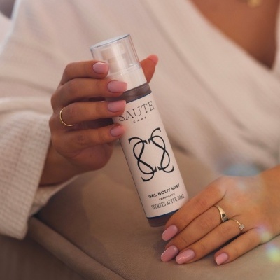 Frasco branco de gel corporal SAUTE CARE com tampa transparente e mão com unhas rosa e anéis