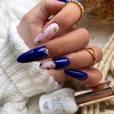 Unhas stiletto azuis e marmorizadas com decoração dourada, anéis dourados e frasco de verniz SAUTE NAILS.