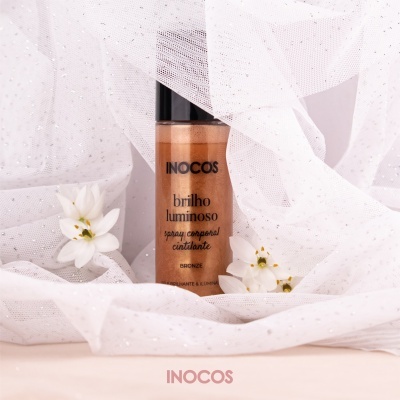 Frasco de spray corporal cintilante INOCOS bronze sobre tecido branco com flores.