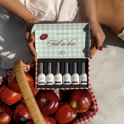 Caixa de vernizes de unhas com padrão xadrez verde e branco e 6 frascos brancos com tampas prateadas sobre uma colcha branca ao lado de uma cesta com maçãs vermelhas.