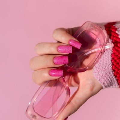 Mão com unhas rosa segurando óculos de sol transparentes cor de rosa.