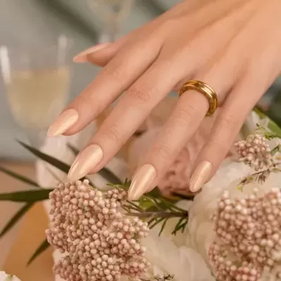 Mão feminina com unhas bege creme usando anel dourado junto a flores brancas e cor de rosa
