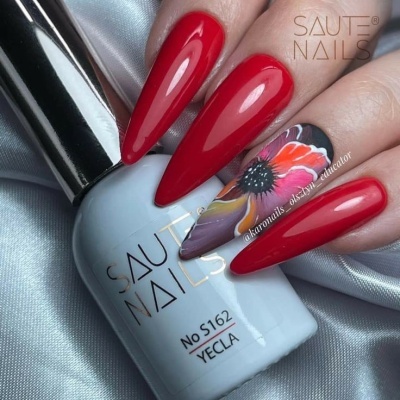 Unhas stiletto vermelhas e uma com decoração floral segurando frasco de verniz SAUT NAILS.
