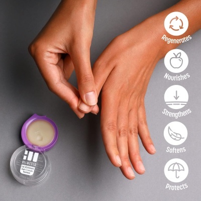 Creme para mãos em embalagem redonda com tampa roxa ao lado de mãos femininas sobre fundo cinza.