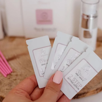 Mão segurando amostras brancas de cosméticos Brown Kross com texto visível, fundo com frascos e caixa em madeira