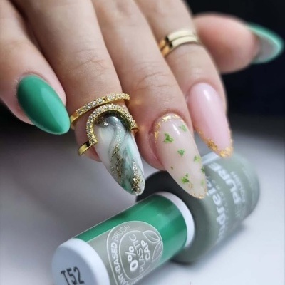 Unhas decoradas com verniz em tons verde e bege, detalhes dourados e folhas, ao lado de frascos de verniz verde e branco.
