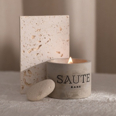 Vela perfumada cinza claro com texto SAUTE CARE, pedra oval e bloco de pedra sobre superfície clara