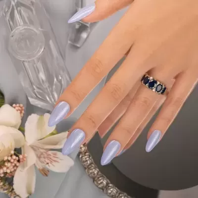 Mão feminina com unhas pintadas de lilás e anel de pedras azuis e brancas, com acessórios ao fundo