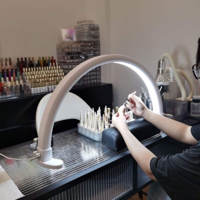 Mesa de manicure com lâmpada branca arqueada, esmaltes e pessoa aplicando unhas postiças