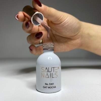 Frasco de verniz para unhas SAUTE NAILS cor OAT MOCHA com mão feminina segurando tampa.
