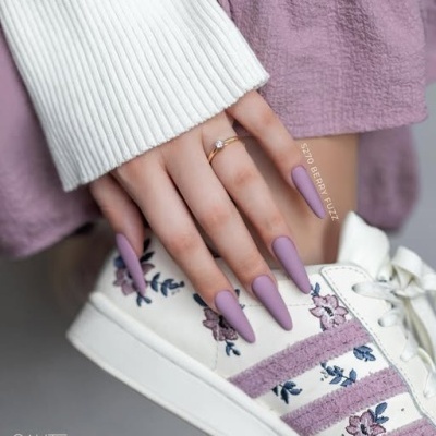 Mãos com unhas lilás fosco segurando ténis branco com riscas e bordados lilás