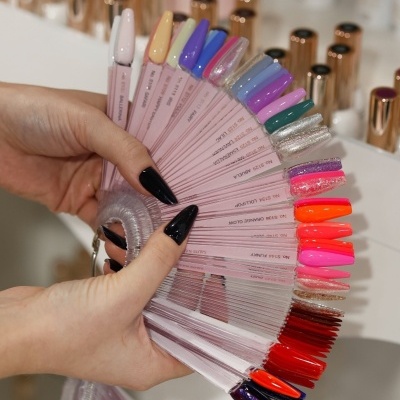 Paleta de amostras de vernizes para unhas em várias cores e acabamentos segurada por mãos com unhas pretas.