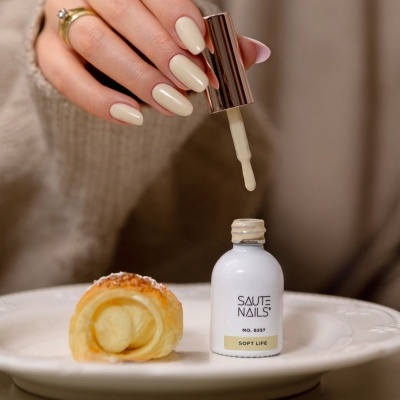 Mãos segurando aplicador de verniz bege claro SAUTE NAILS ao lado de pastelaria num prato branco