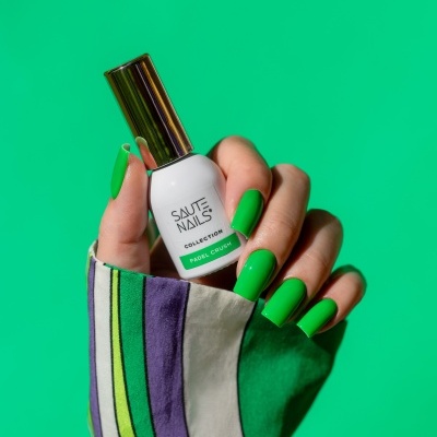 Frasco de verniz de unhas branco e dourado com texto PADEL CRUSH, fundo verde e mão com unhas verdes.